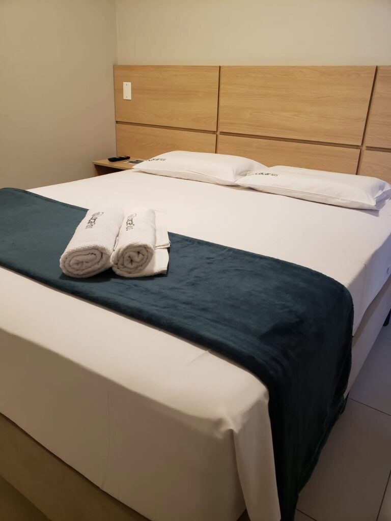 Cama de casal com lençóis brancos, manta azul-escura e duas toalhas enroladas