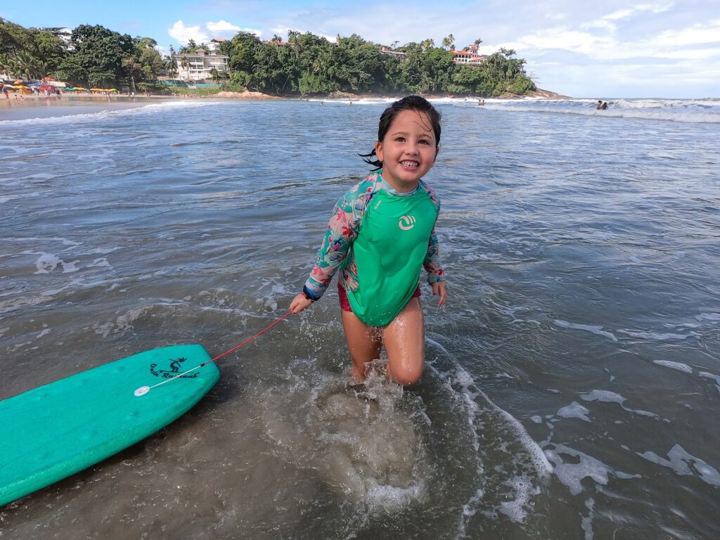 Menina sorridente segurando prancha de bodyboard verde na beira do mar da Praia do Tenório