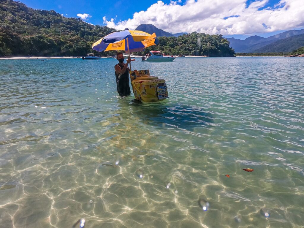 Vendedor ambulante com guarda-sol e caixa térmica dentro do mar raso e cristalino de uma praia de Ubatuba, com serra ao fundo