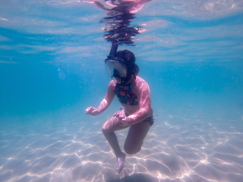 Pessoa fazendo snorkel em água azul-clara e transparente próxima ao fundo arenoso de uma praia de Ubatuba
