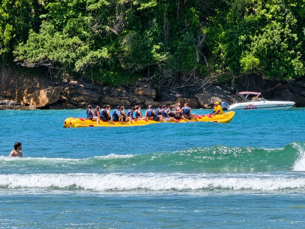 Banana boat amarelo com grupo de pessoas de colete passando perto de um costão coberto de vegetação