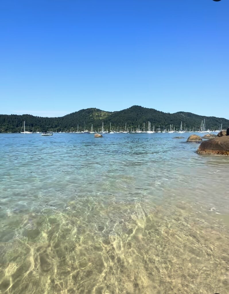 Água rasa e transparente em praia de Ubatuba com montanhas ao fundo e veleiros no horizonte