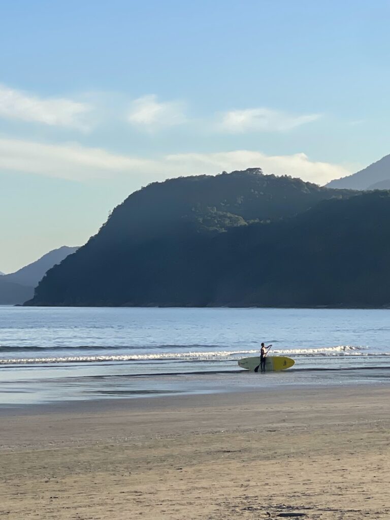 Surfista com prancha na areia de praia ampla em Ubatuba, com morro ao fundo