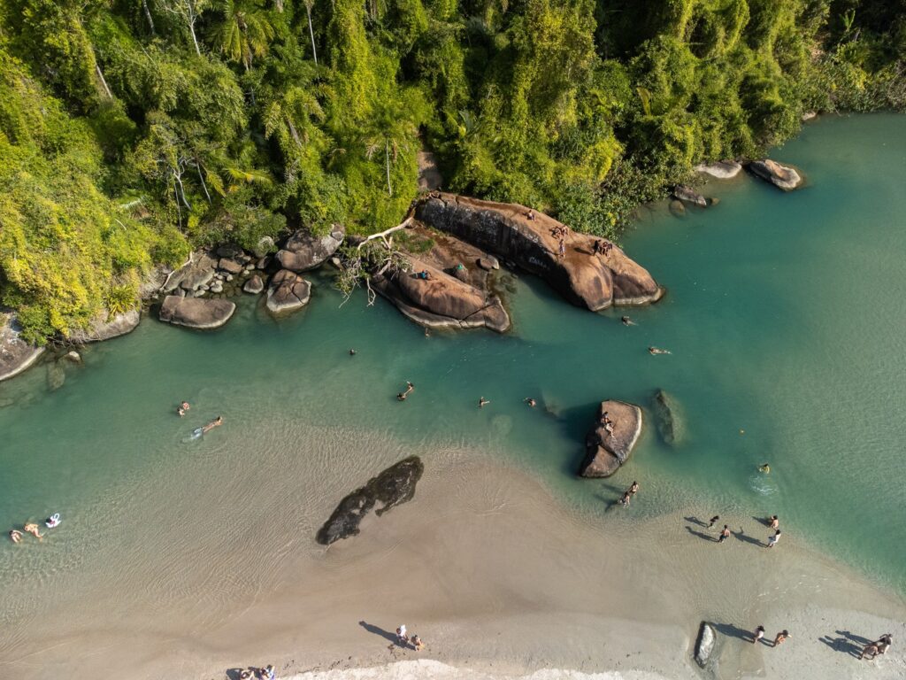 Vista aérea do rio Itamambuca encontrando a praia em Ubatuba, com água verde, pedras e banhistas.