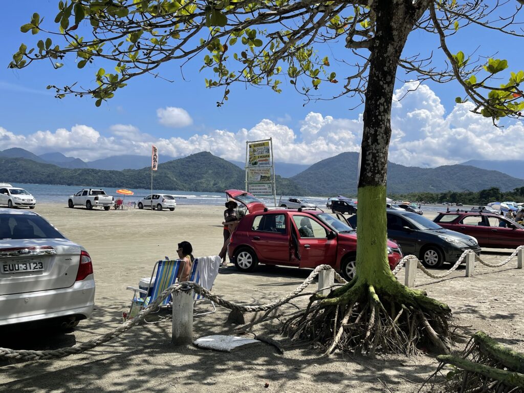 Carros estacionados na faixa de areia de Ubatumirim com mar e montanhas ao fundo